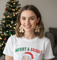 Woman wearing gingerbread couple drop earrings, smiling in front of a Christmas tree