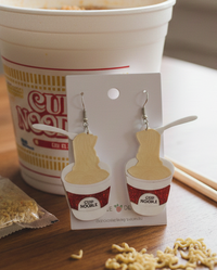 Acrylic cup noodle drop earrings on a card, showing spoon and 'Cup Noodle' label detail against a real noodle cup