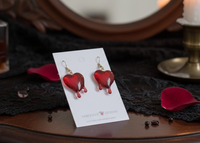 Pair of red dripping heart drop earrings on card against a dark gothic dresser scene with candle