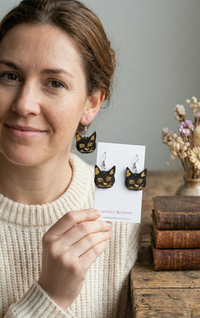 Woman wearing one black cat face earring with gold detail, second earring shown on card
