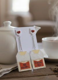 Tea bag drop earrings with red heart studs on a card, reading Be Happy and Drink Tea, styled with a teapot