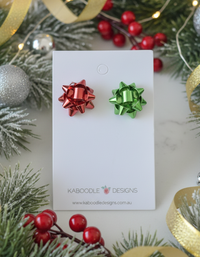 Red and green metallic gift bow stud earrings on a Kaboodle Designs card surrounded by Christmas greenery