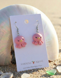 Pink resin octopus dangle earrings with cartoon eyes on Kaboodle Designs card, shown against a shell