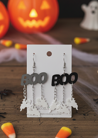 Black and grey acrylic BOO earrings with dangling ghost charms on display card, jack-o'-lanterns behind