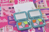 Pair of blue makeup palette earrings laid flat showing mirror and coloured eyeshadow squares with brush charm