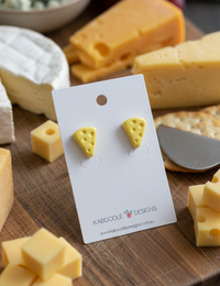 Yellow resin cheese wedge stud earrings with visible holes, displayed on a card among cheese blocks