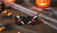 Pair of black acrylic knife-through-heart earrings laid flat on wooden surface with Halloween spiders and candy corn