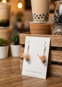 Resin milkshake drop earrings displayed on branded card in front of an iced boba drink