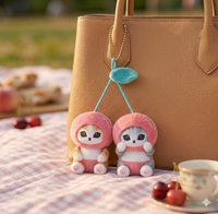 Two plush cherry-costume cat keychains hanging from a tan bag, cherries and teacup in foreground