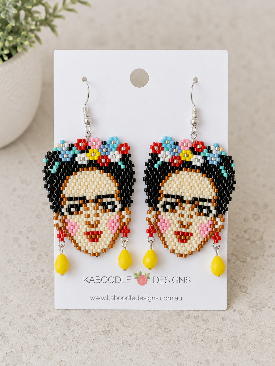 Seed bead drop earrings shaped like a face with floral crown and yellow bead dangle, on card