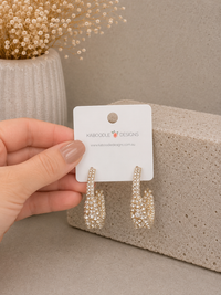 Gold-toned diamante hoop earrings held in hand against a stone block and vase of dried flowers