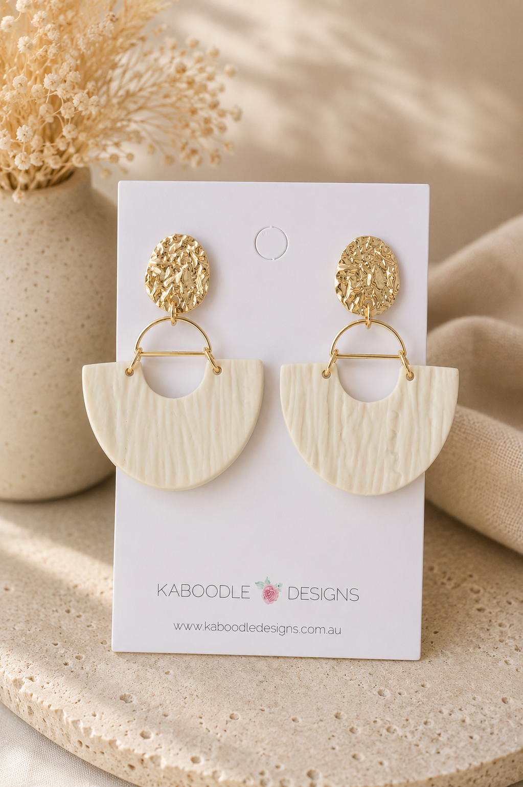 Beige clay half-moon drop earrings with textured finish on gold hammered studs, shown on card