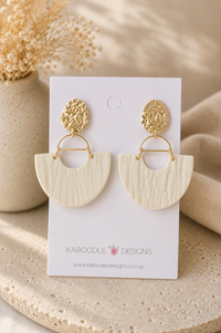 Beige clay half-moon drop earrings with textured finish on gold hammered studs, shown on card