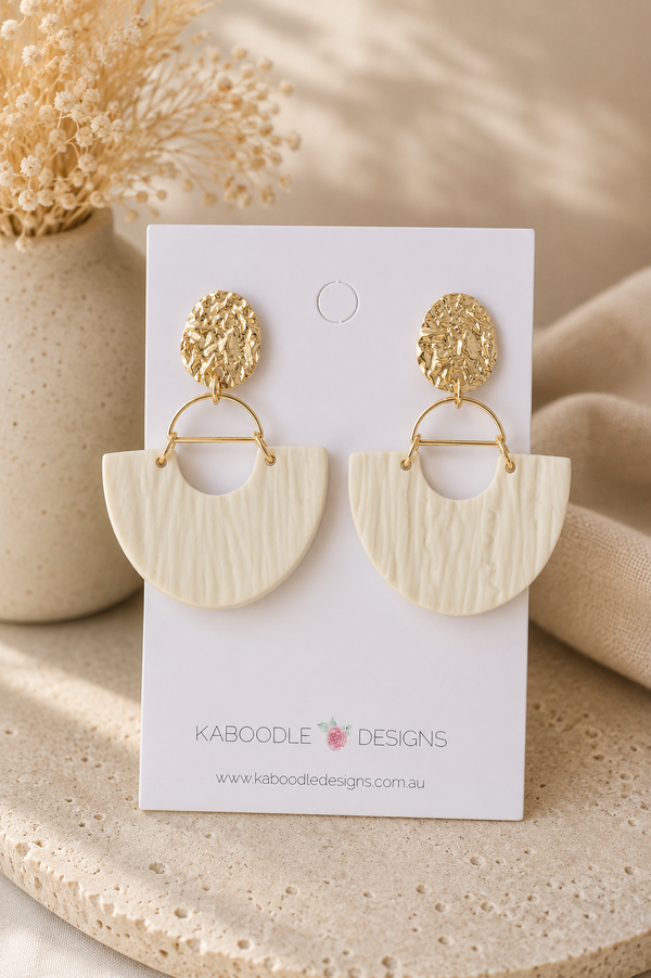 Beige clay half-moon drop earrings with textured finish on gold hammered studs, shown on card