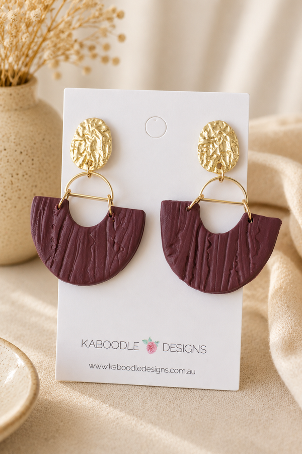Burgundy clay fan-shaped drop earrings with hammered gold-tone studs on display card