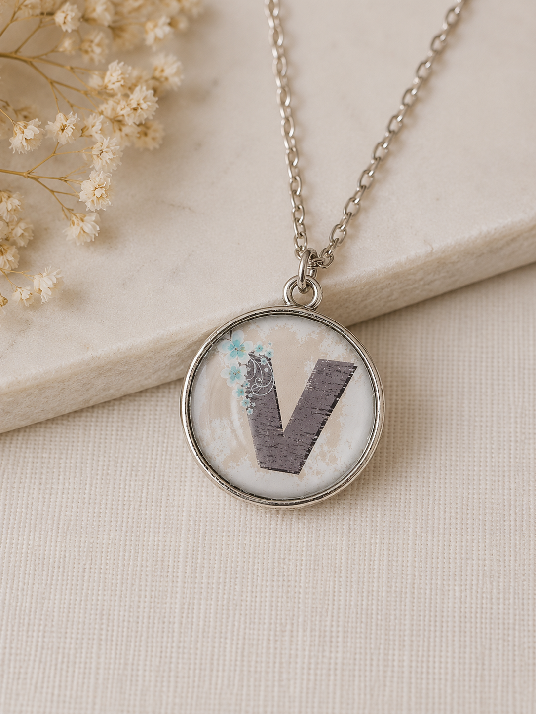 Round silver-tone pendant with letter V and blue floral detail, resting on stone tile