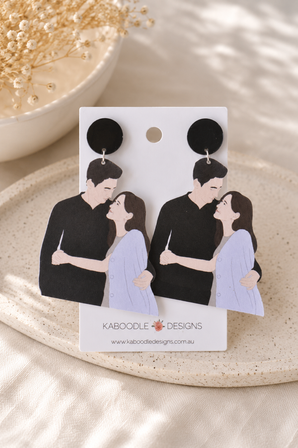 Pair of Kdrama couple drop earrings on card, illustrated man and woman embracing under black stud