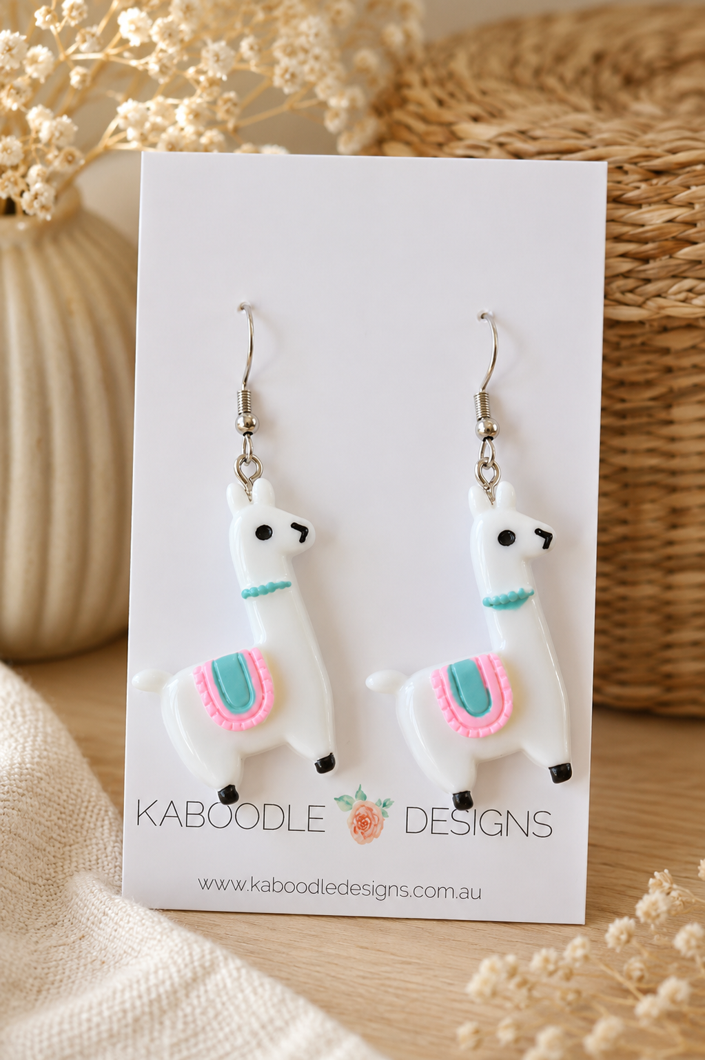 Pair of white resin llama drop earrings with pink and teal saddle detail on card