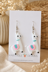 Pair of white resin llama drop earrings with pink and teal saddle detail on card
