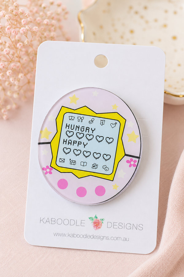 Round acrylic Tamagotchi brooch in pink and yellow with pixel heart icons, shown on card