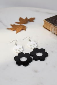Close-up of black and white skull-and-flower earrings on a marble surface with autumn leaves