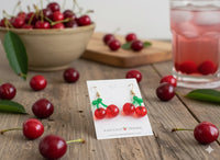 Close-up of cherry earrings on card surrounded by fresh cherries and a pink drink