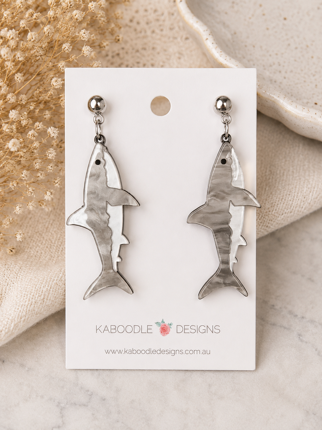 Pair of marbled grey and white shark-shaped dangle earrings with silver ball-top studs, displayed on a card.