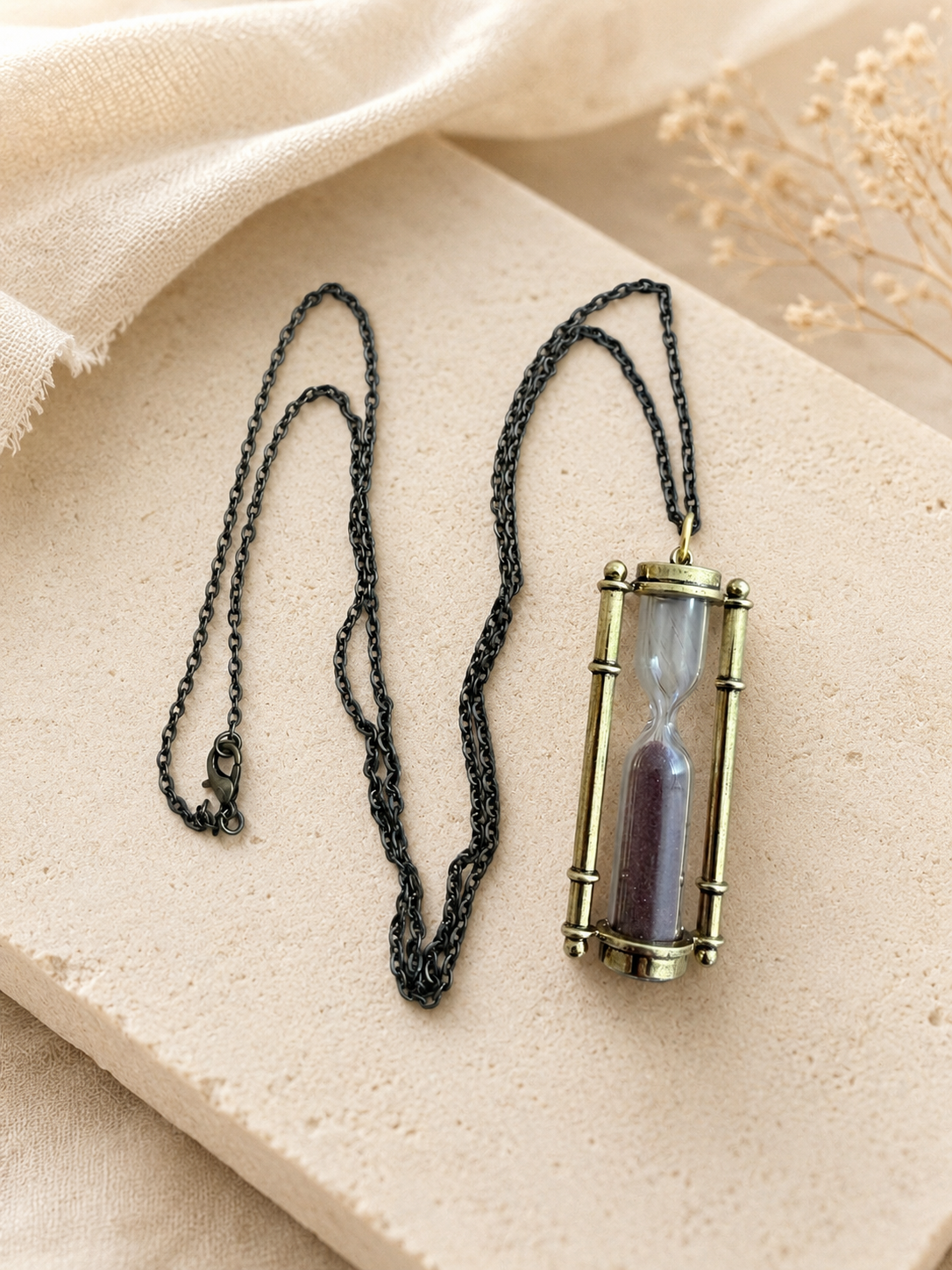 Hourglass pendant necklace with dark sand inside a rounded frame on a dark chain, laid on a stone tile.