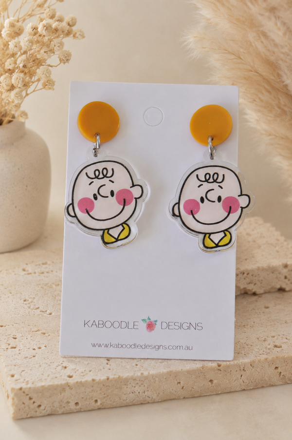 Pair of drop earrings with mustard yellow circle studs and smiling cartoon boy face charms with rosy cheeks and a yellow