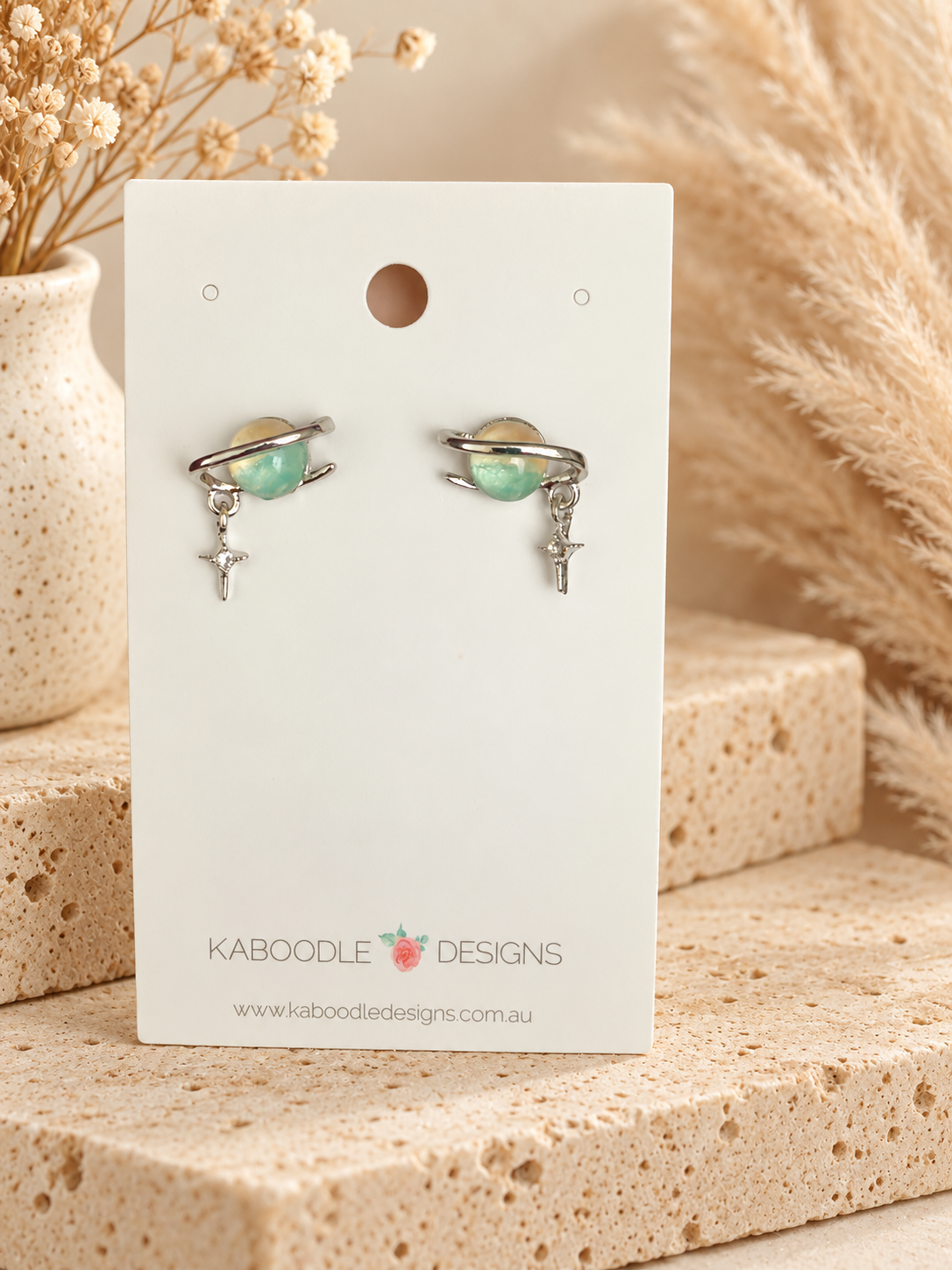 Pair of Saturn-shaped stud earrings with green planet, silver ring and star charm on a display card