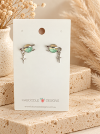 Pair of Saturn-shaped stud earrings with green planet, silver ring and star charm on a display card