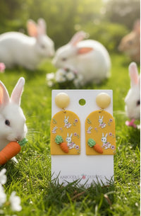 Mustard bunny carrot earrings displayed outdoors on grass with real rabbits in background