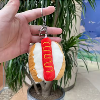 Plush hot dog keychain with mustard zigzag detail held in hand against greenery