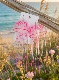 Pair of pink jellyfish dangle earrings with translucent domes and long wavy tentacles, hung on driftwood by the beach