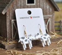 Pair of resin cow drop earrings with black spots on a Kaboodle Designs card against a barn backdrop