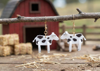 Two cow earrings hanging from a branch, showing full 3D body and legs against a barn scene