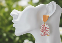 Close-up of geometric flower earring worn on ear, showing rust-red floral line design