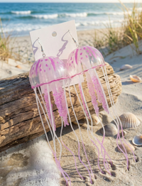 Pair of purple jellyfish acrylic dangle earrings on display card against beach driftwood and shells