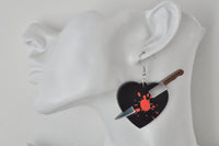 Close-up of broken heart knife earring worn on ear, showing brown knife handle and silver blade detail against black heart