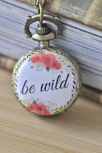 Bronze pocket watch necklace with pink rose wreath and be wild quote design, resting on a book