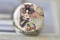 Close-up of pocket watch pendant lid featuring cat playing violin artwork with birds
