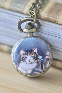 Brass pocket watch necklace with vintage kitten artwork on a wooden shelf beside books