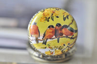 Close-up of the four birds artwork on the pocket watch pendant lid, showing orange robins amid yellow blossoms