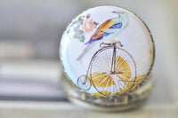 Close-up of penny farthing and bird artwork on round pocket watch pendant