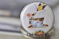 Close up of tea cup and songbird artwork on round pocket watch pendant