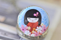 Close-up of kokeshi doll pendant artwork with butterfly hair clip and pink floral border