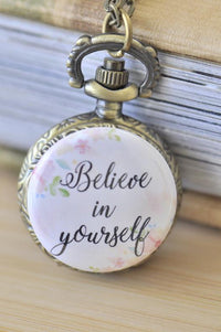 Bronze pocket watch necklace with floral "Believe in yourself" design resting against a stack of books