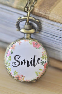 Antique bronze pocket watch necklace with floral 'Smile' design pendant resting against a book