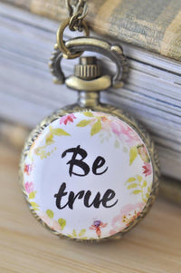 Close up of pocket watch necklace with floral wreath and Be true script on the face