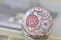 Close up of floral swirl pattern in pink, lilac and cream on antique bronze pocket watch pendant